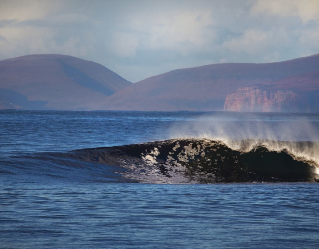 A surfer carves through a massive right-hander at Thurso East against a backdrop of the Scottish Highlands